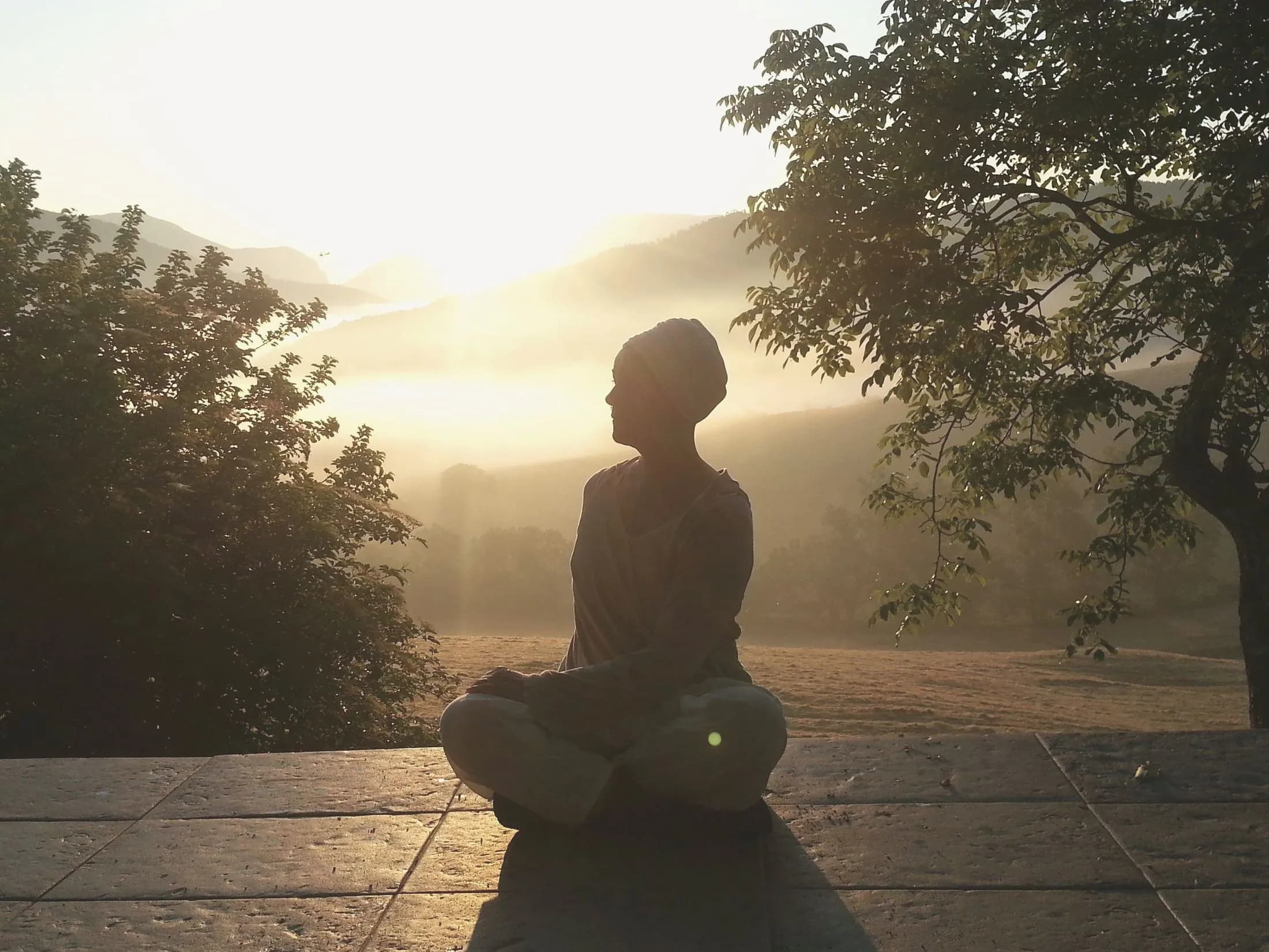 Jogini istuu meditaatiossa kivitasanteella auringonnousun aikaan, taustalla usvaiset kukkulat.